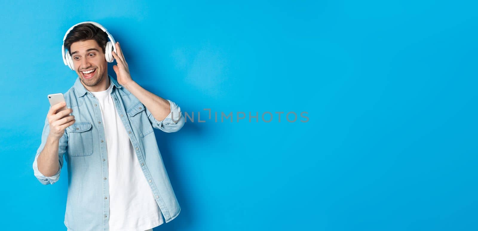 Happy man listening to music in headphones and reading message on smartphone, smiling excited, standing against blue background.
