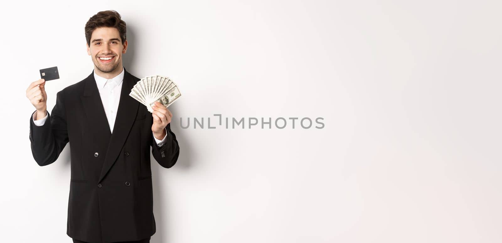 Portrait of attractive businessman in black suit, showing money and credit card, smiling pleased, standing against white background by Benzoix