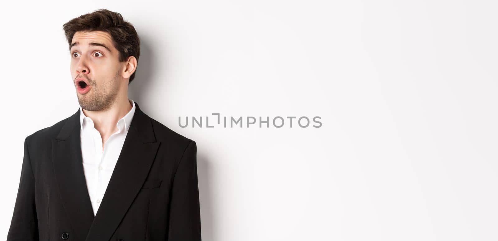 Close-up of impressed guy in trendy suit, open mouth and looking left wondered, standing against white background.