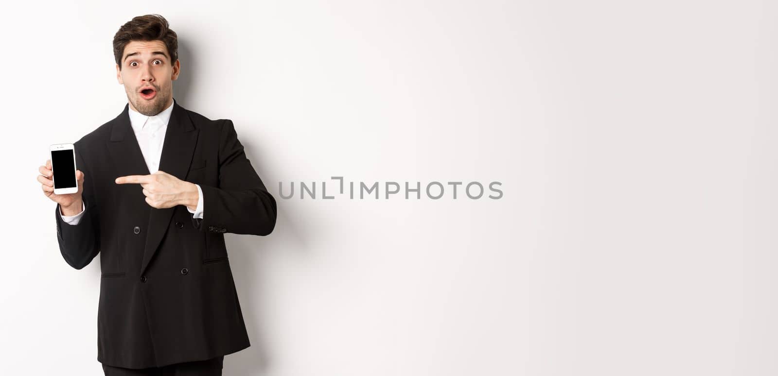 Portrait of attractive businessman in black suit, looking surprised and pointing finger at smartphone sreen, standing over white background.