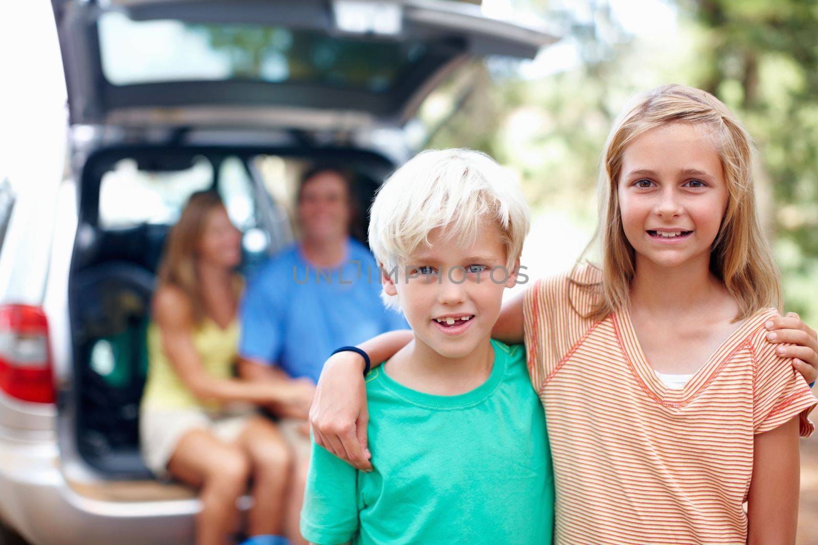 Brother and sister standing with arms around and smiling. Brother and sister standing with arms around and parents sitting in the back of a car. by YuriArcurs