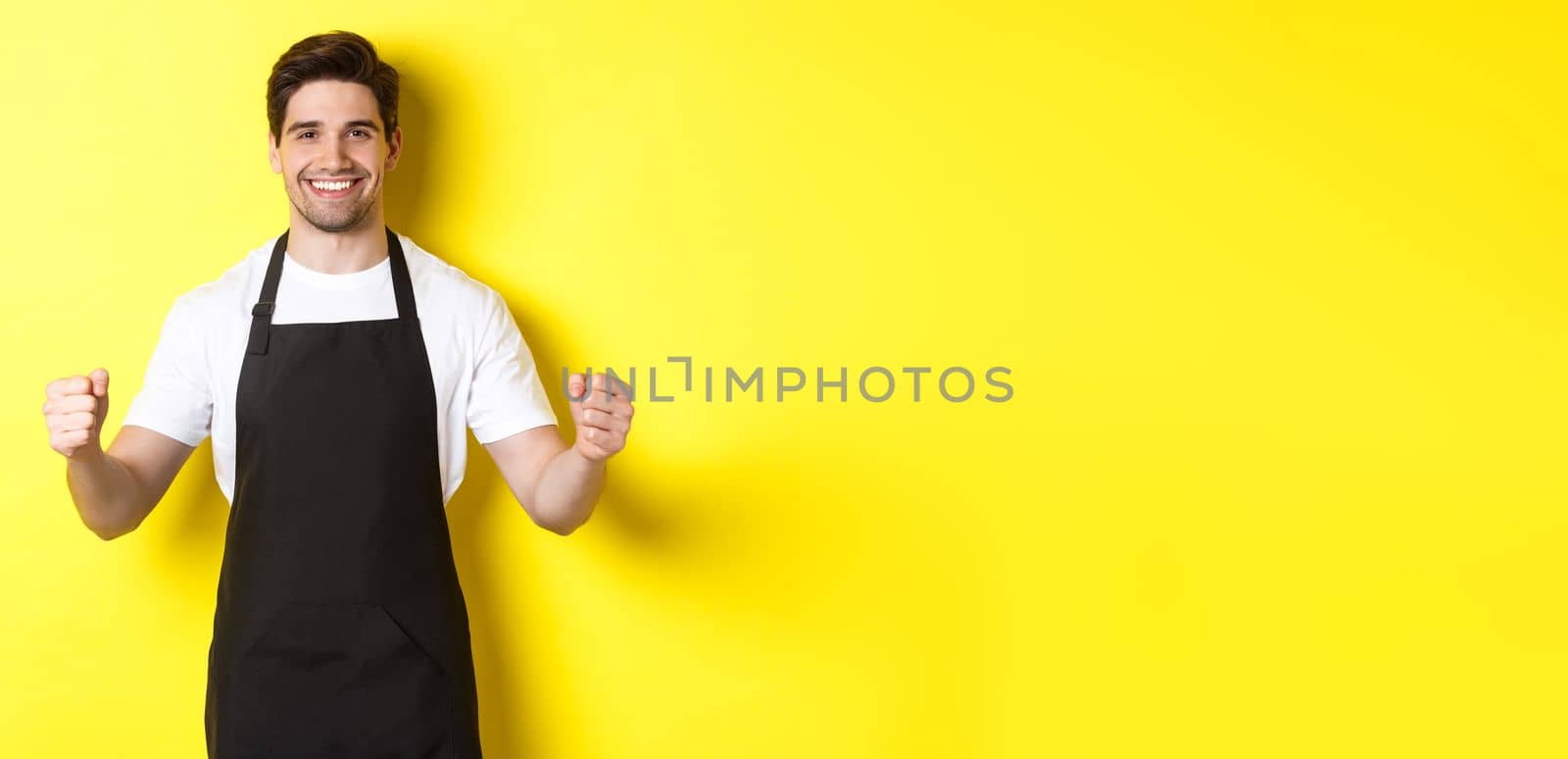 Friendly coffee shop waiter standing with raised hands, place for your sign or logo, standing over yellow background by Benzoix