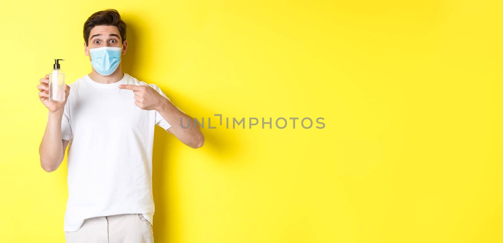 Concept of covid-19, quarantine and lifestyle. Excited guy in medical mask showing good hand sanitizer, pointing finger at antiseptic, standing over yellow background by Benzoix