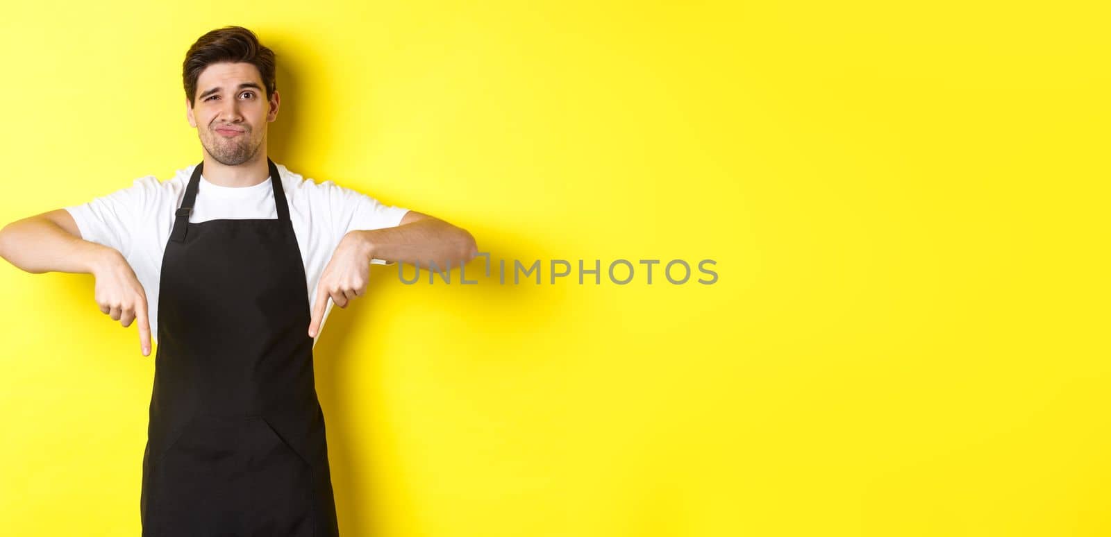 Skeptical barista in black apron pointing fingers down at bad product, looking displeased and unamused, standing over yellow background.