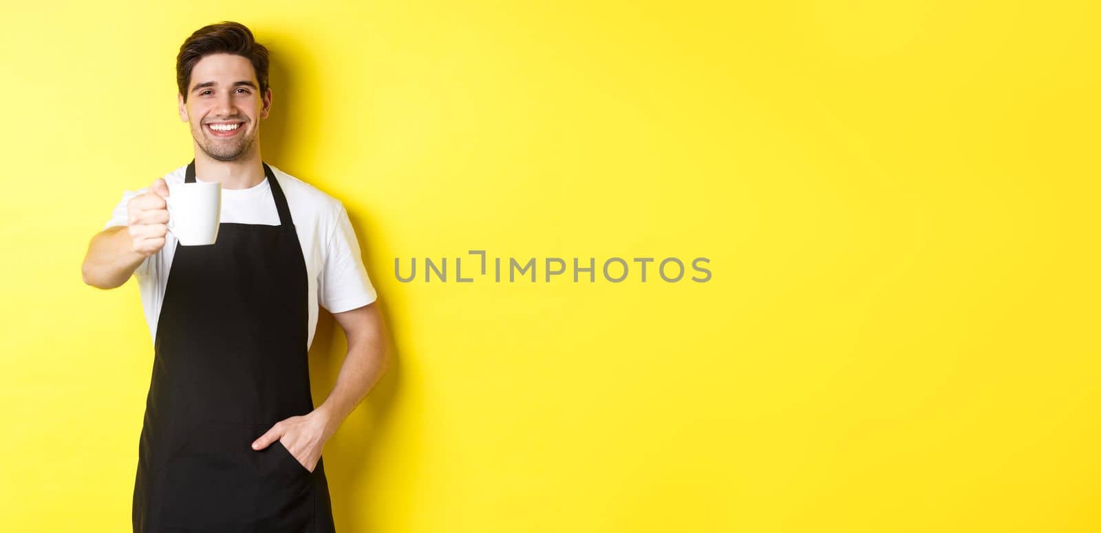 Handsome barista in black apron giving you cup of coffee and smiling, standing over yellow background by Benzoix