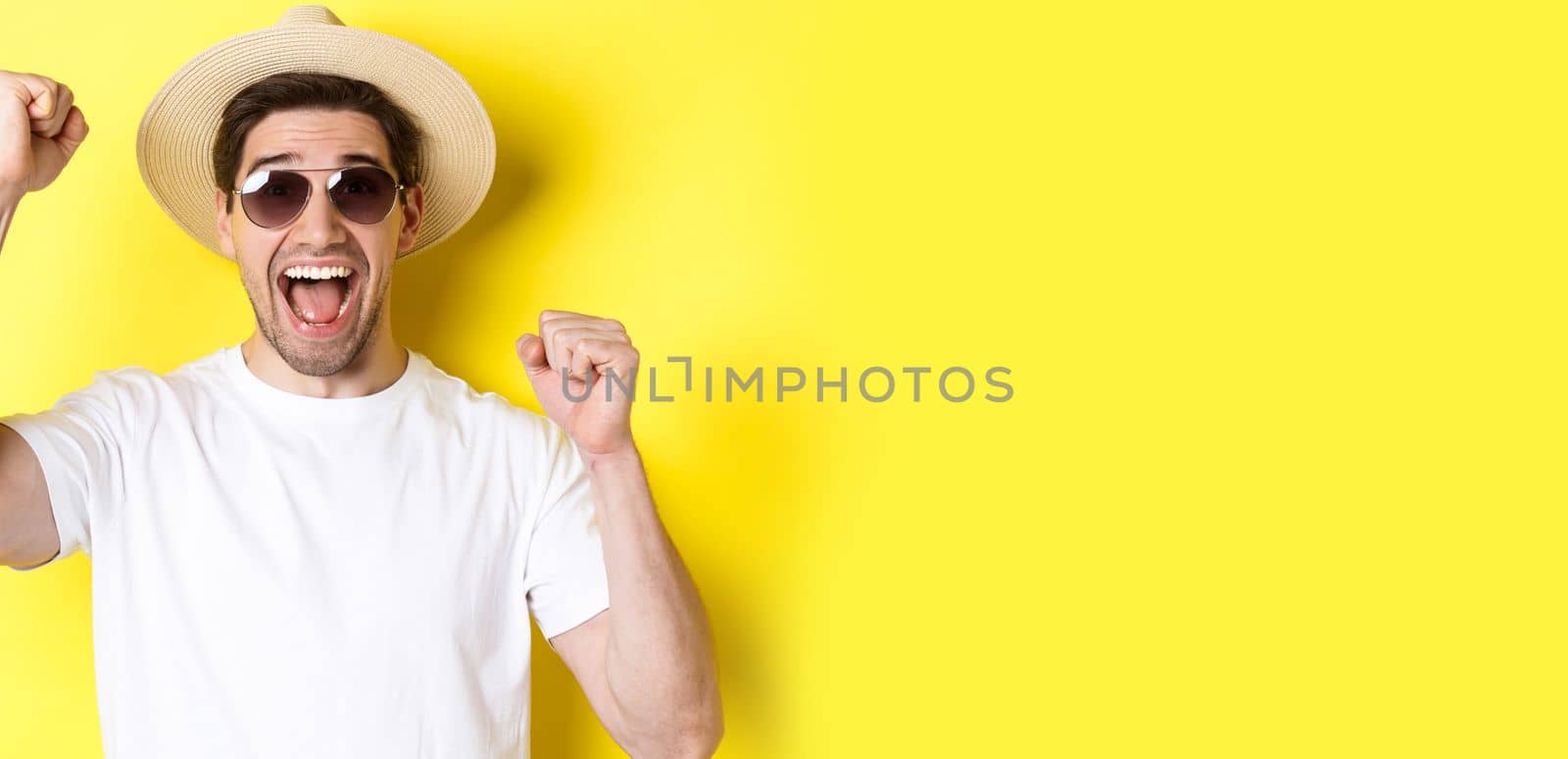 Concept of tourism and holidays. Happy male tourist celebrating his vacation, raising hands up and shouting for joy, wearing sunglasses with straw hat.