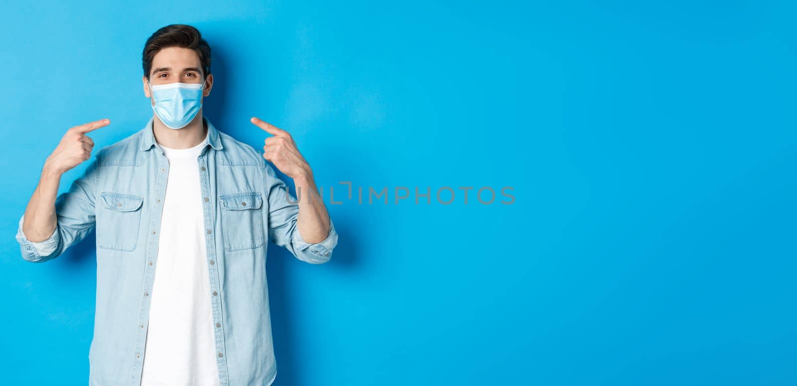 Concept of coronavirus, quarantine and social distancing. Handsome man pointing at medical mask and smiling, protection from virus spread during pandemic, blue background.