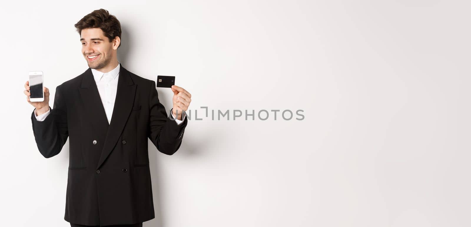 Handsome successful businessman, looking at smartphone screen and showing credit card, standing in black suit against white background by Benzoix