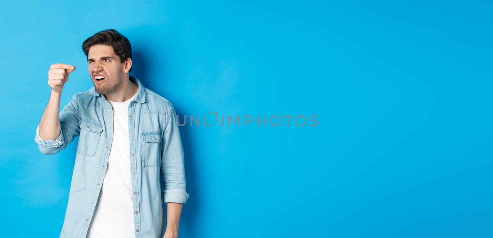 Confused guy holding something tiny in fingers and looking at it puzzled, standing over blue background by Benzoix