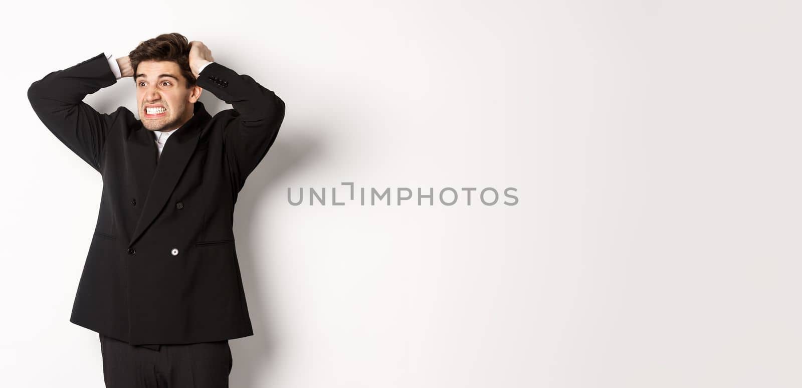 Image of frustrated and angry businessman in black suit, ripping hair on head and grimacing mad, looking left at disaster, standing tensed against white background.