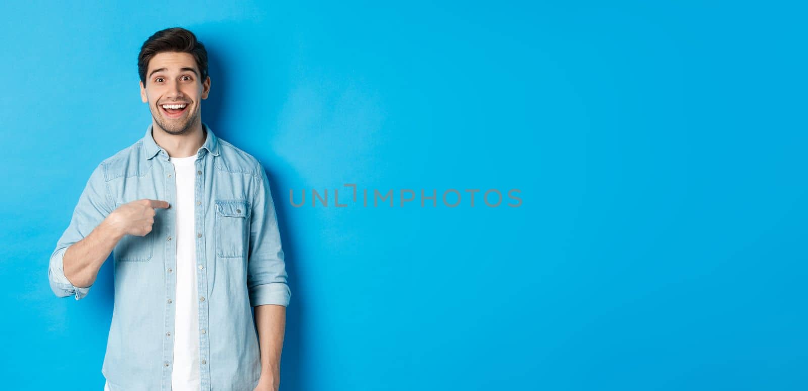 Happy and surprised man pointing at himself, smiling pleased, standing against blue background by Benzoix