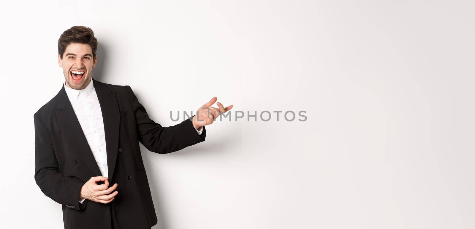 Portrait of happy man dancing at party, playing on invisible guitar and laughing, standing in black suit against white background.