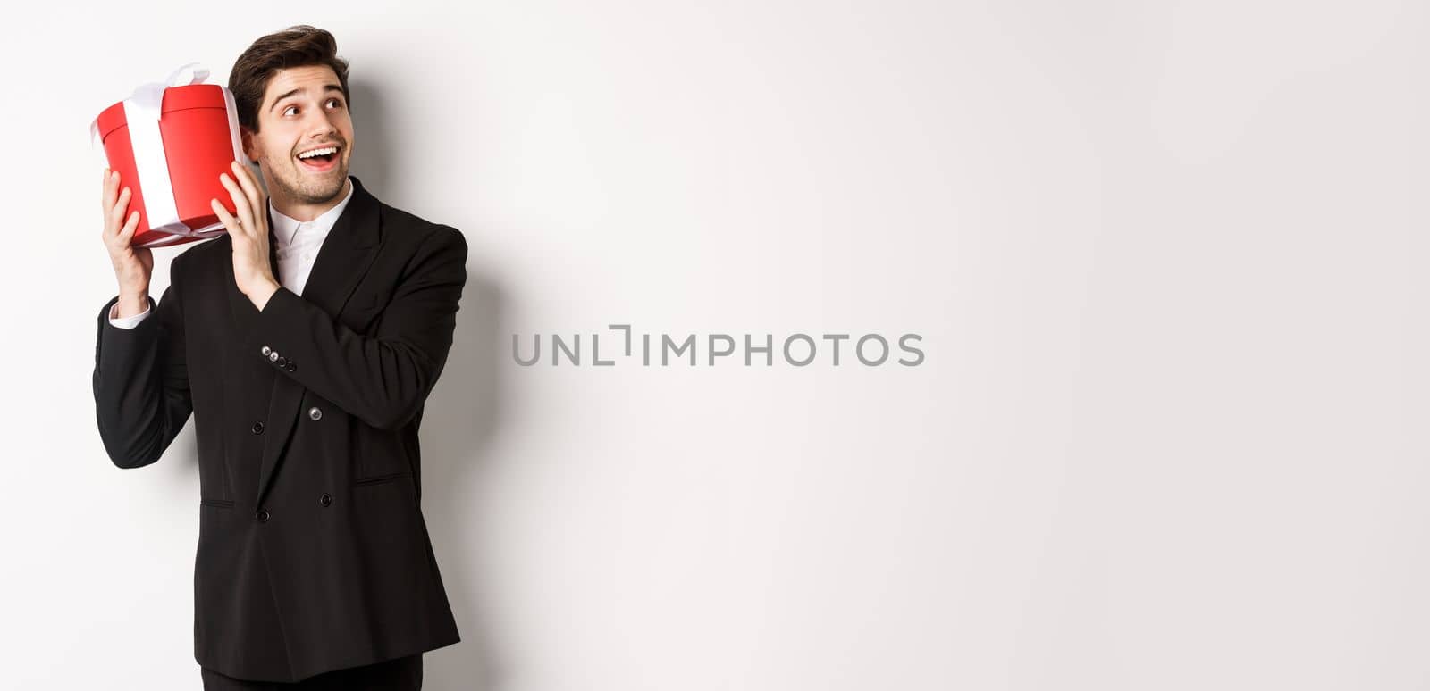 Concept of christmas holidays, celebration and lifestyle. Image of attractive man in black suit, shaking present to guess what inside, enjoying new year, standing against white background by Benzoix