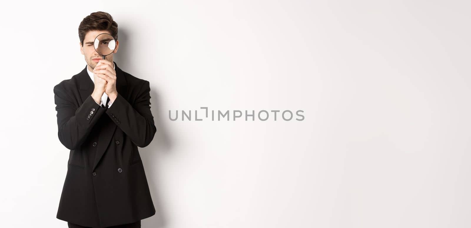 Image of serious businessman in black trendy suit, looking through magnifying glass, looking for employees, standing against white background.