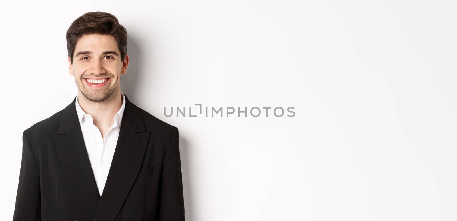 Close-up of handsome young businessman in trendy suit smiling, standing against white background.