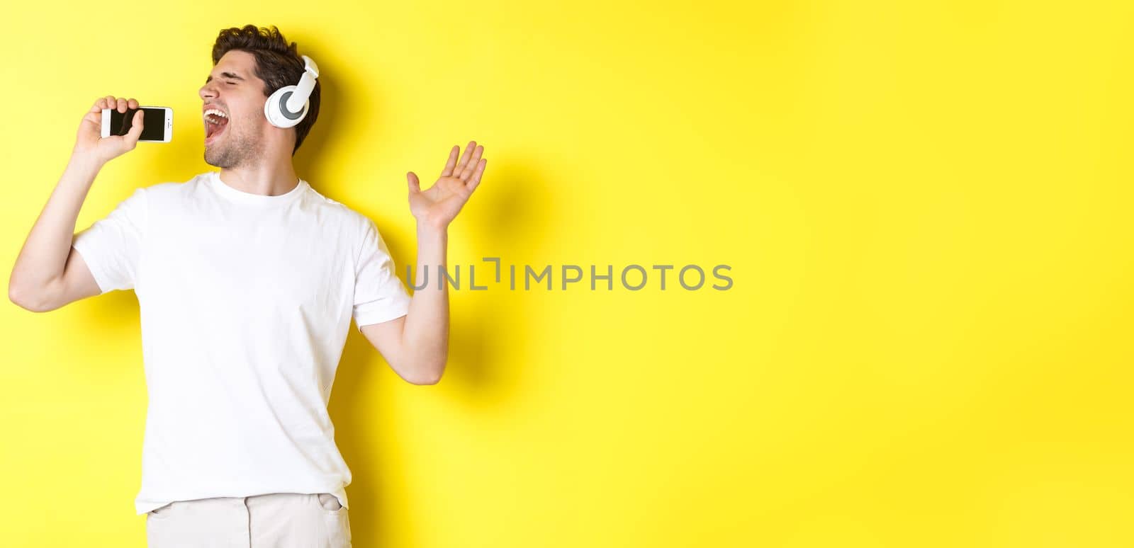 Happy guy playing karaoke app in headphones, singing into smartphone microphone, standing over yellow background.