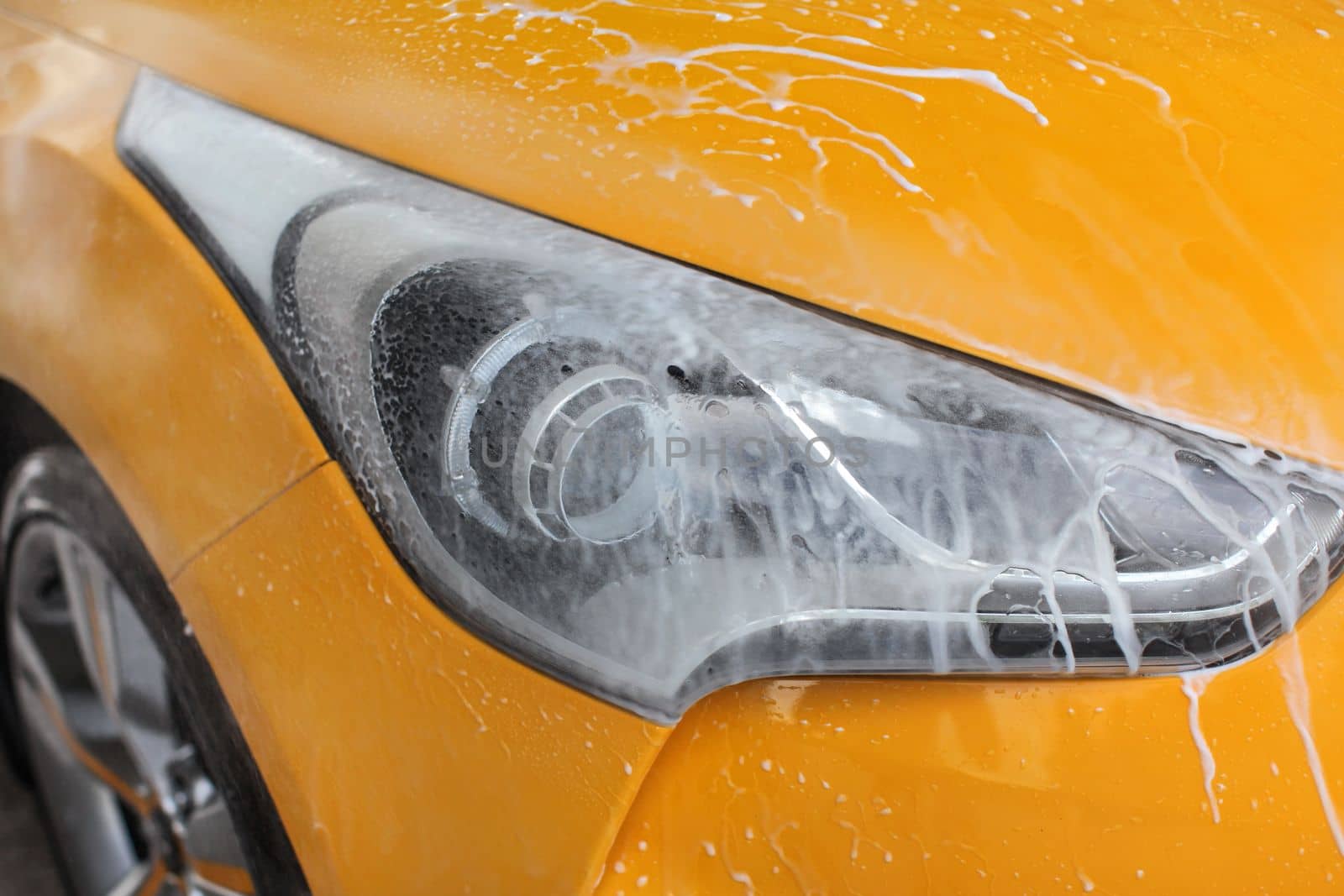 Front light of yellow car washed in carwash, jet water spraying, white shampoo and foam flowing on surface.