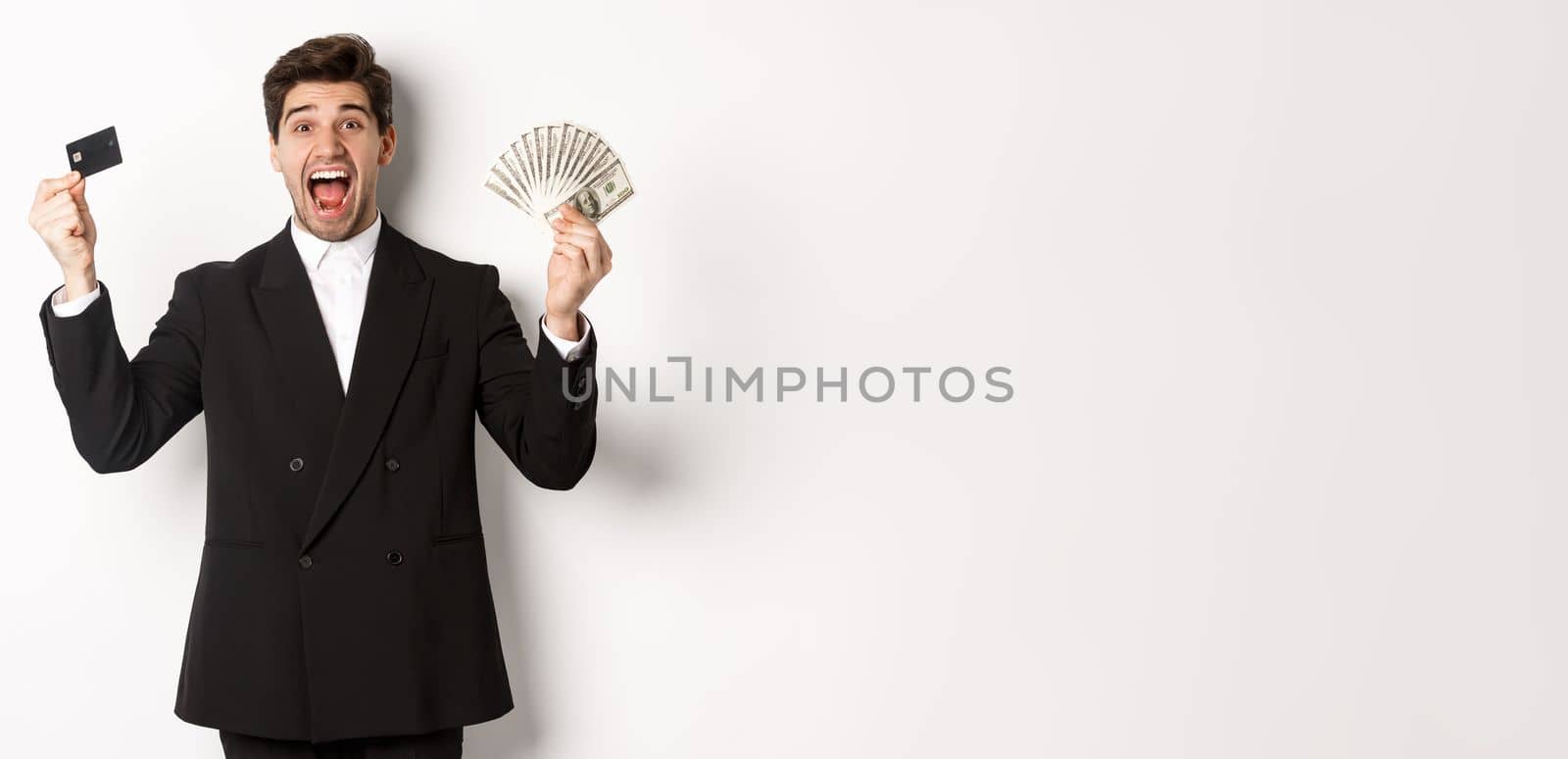 Portrait of handsome businessman in black suit, showing credit card and money, shouting for joy and excitement, standing against white background.