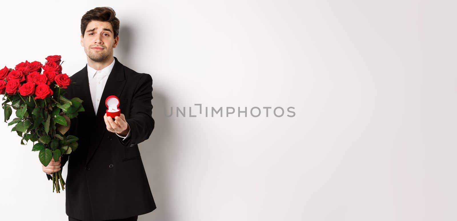 Handsome smiling man in black suit, holding roses and engagement ring, making a proposal to marry him, standing against white background by Benzoix