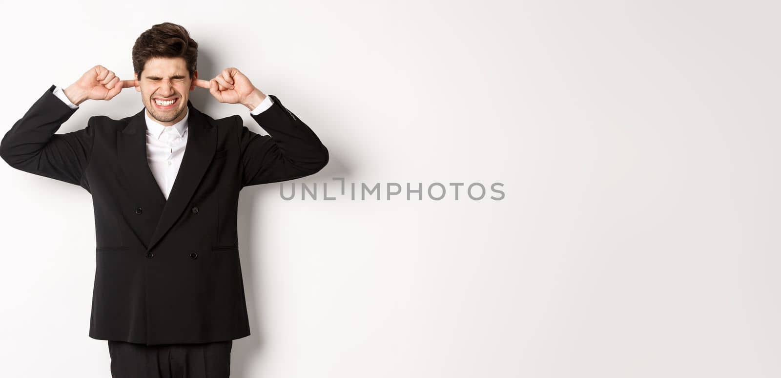 Portrait of annoyed and bothered businessman in black suit, shut ears, feeling uncomfortable, complaining loud noise, standing against white background.