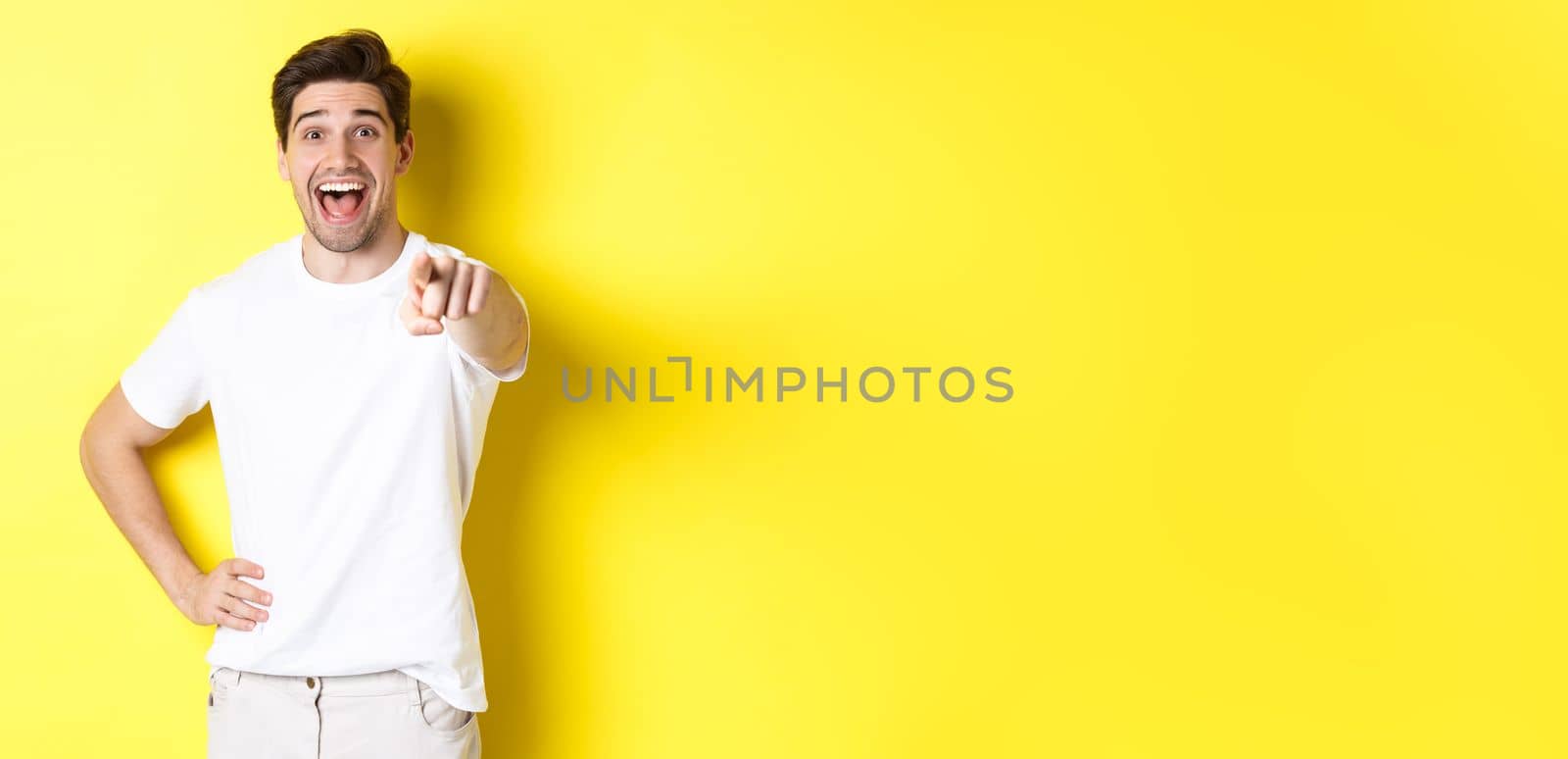 Happy guy pointing finger at camera and laughing, check out something, standing over yellow background.