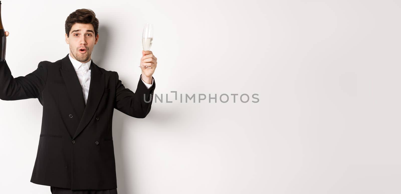 Concept of holidays, party and celebration. Image of handsome man in stylish suit, dancing with bottle of champagne, drinking on new year, standing over white background by Benzoix