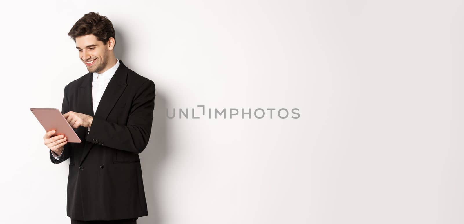 Portrait of attractive man in trendy suit, looking at digital tablet and smiling, shopping online, standing over white background.