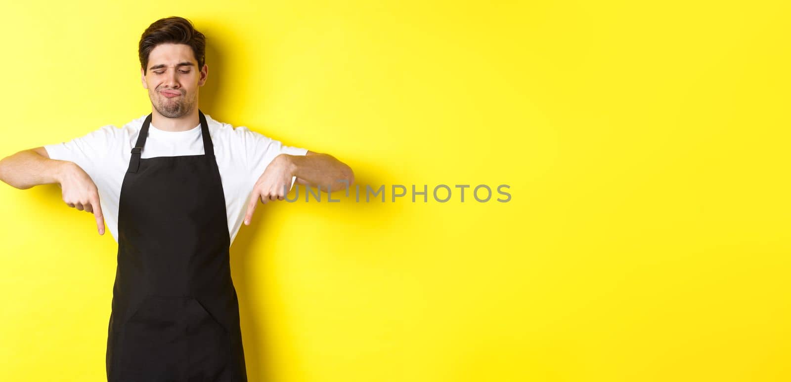 Doubtful barista complaining, pointing fingers down and grimacing displeased, standing in black apron against yellow background by Benzoix