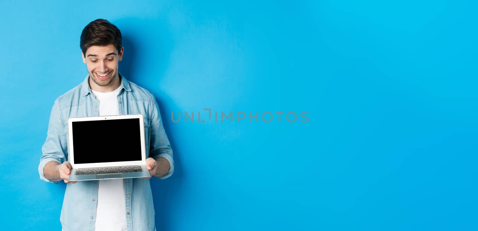 Excited handsome man looking at screen, showing promo offer on computer screen, smiling amazed, standing over blue background.