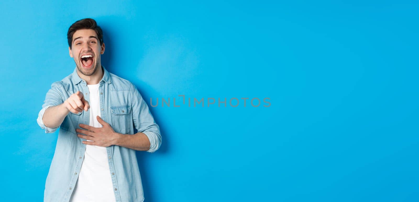 Adult man in casual outfit laughing out loud and pointing at you, looking at something funny, chuckling while standing against blue background.