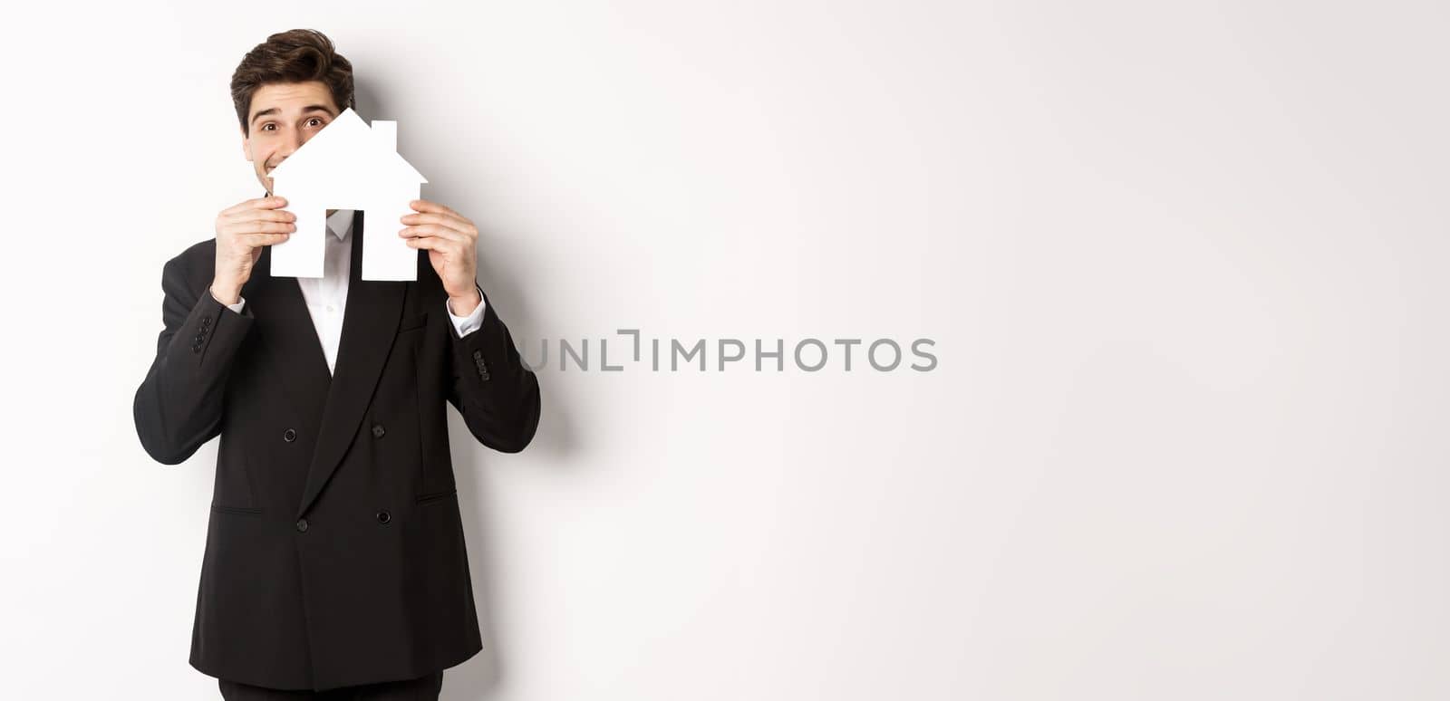 Image of handsome broker in black suit, showing house maket and smiling, selling homes, standing against white background.