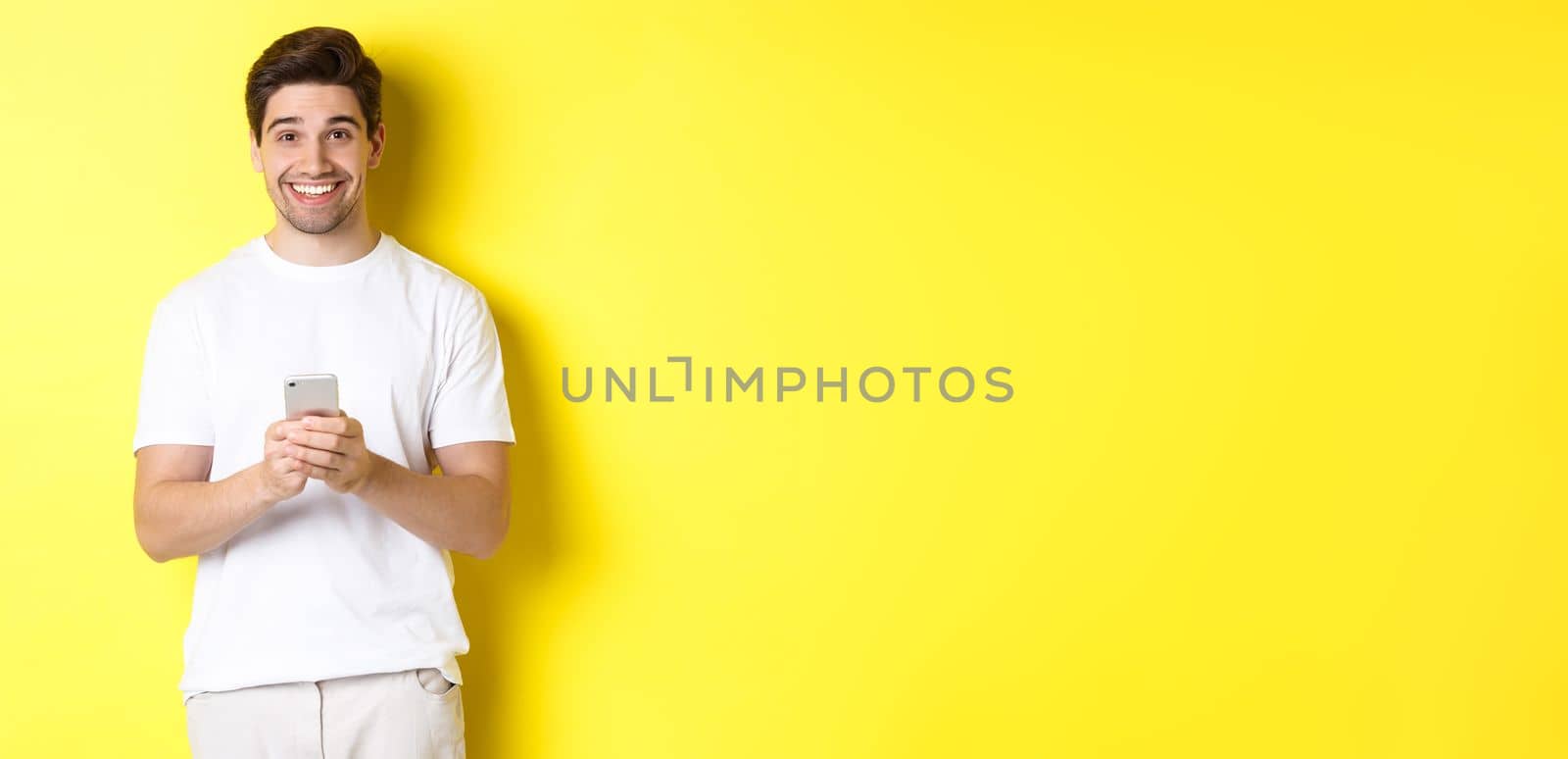 Man smiling and looking happy after reading promo offer on smartphone, standing against yellow background in white t-shirt.