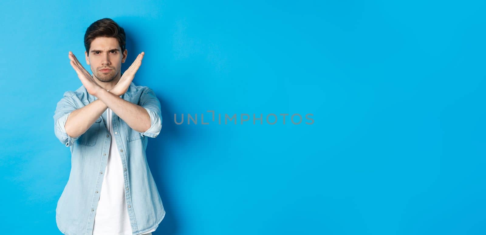 Displeased man frowning and saying no, making cross sign, tell to stop, standing against blue background.