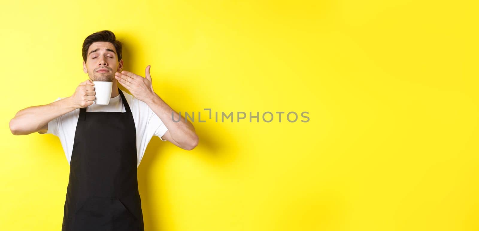 Barista enjoying smell of coffee in mug, standing pleased with eyes closed, wearing black apron.