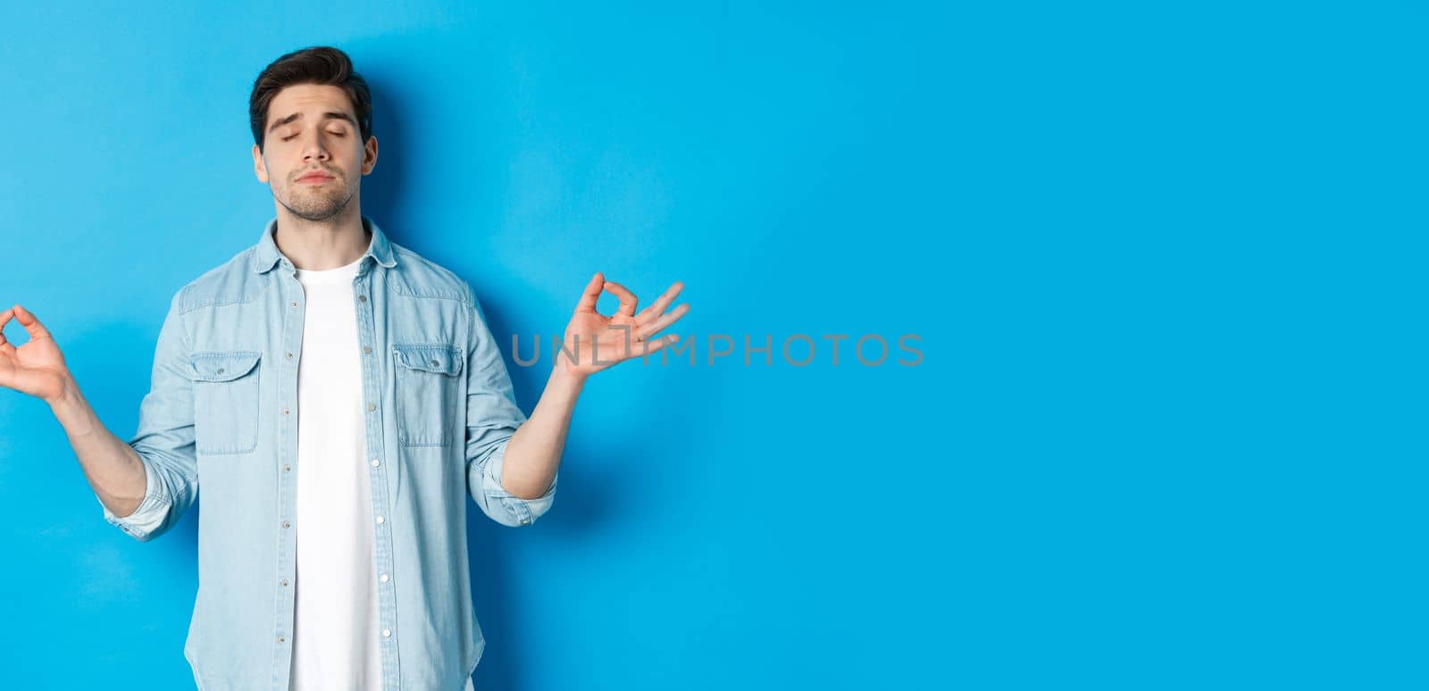 Calm man with closed eyes meditating, holding hands sideways and do yoga breathing exercises, standing against blue background by Benzoix