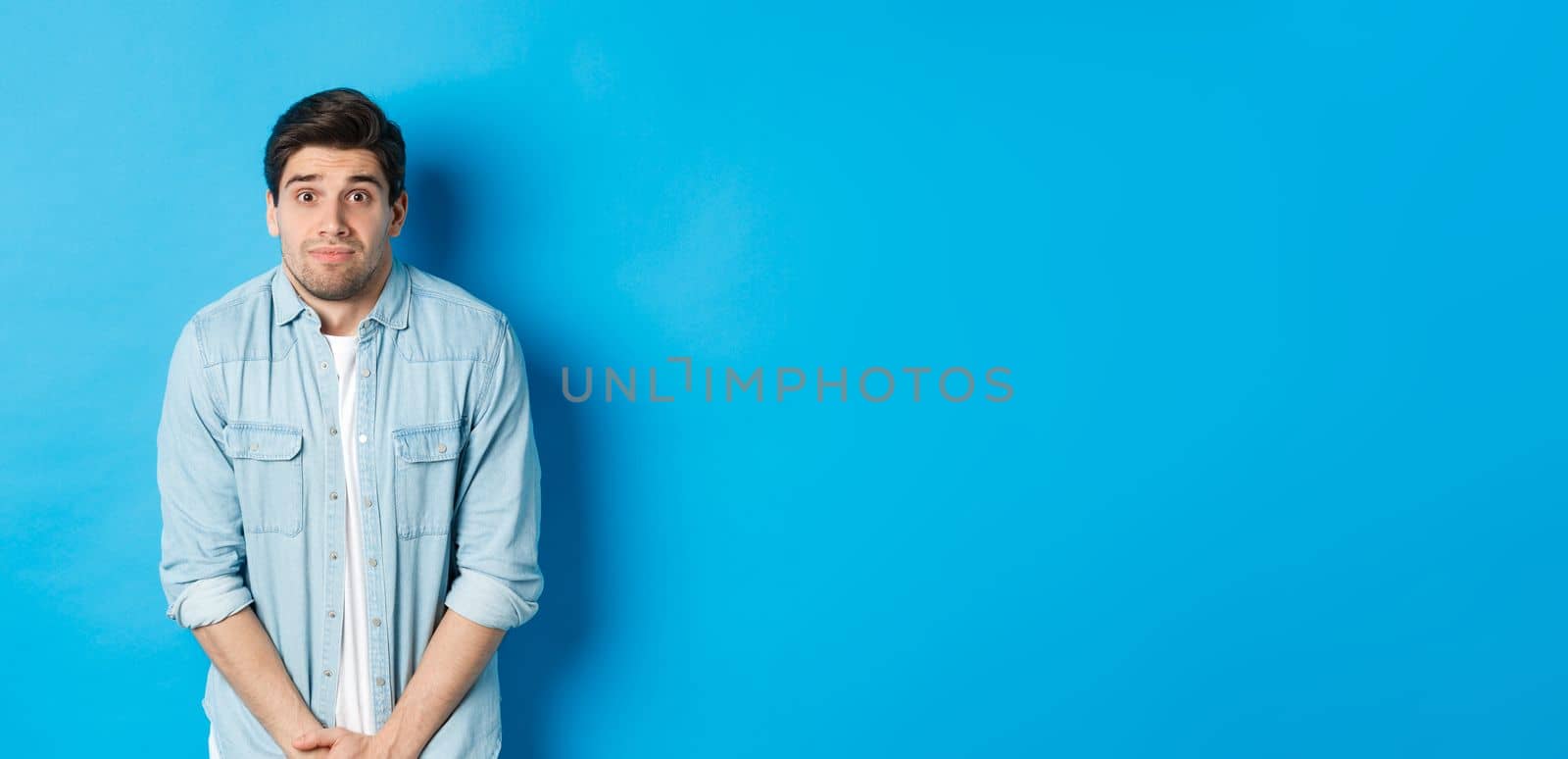 Embarrassed guy want to pee, waiting in line for toilet, standing against blue background.