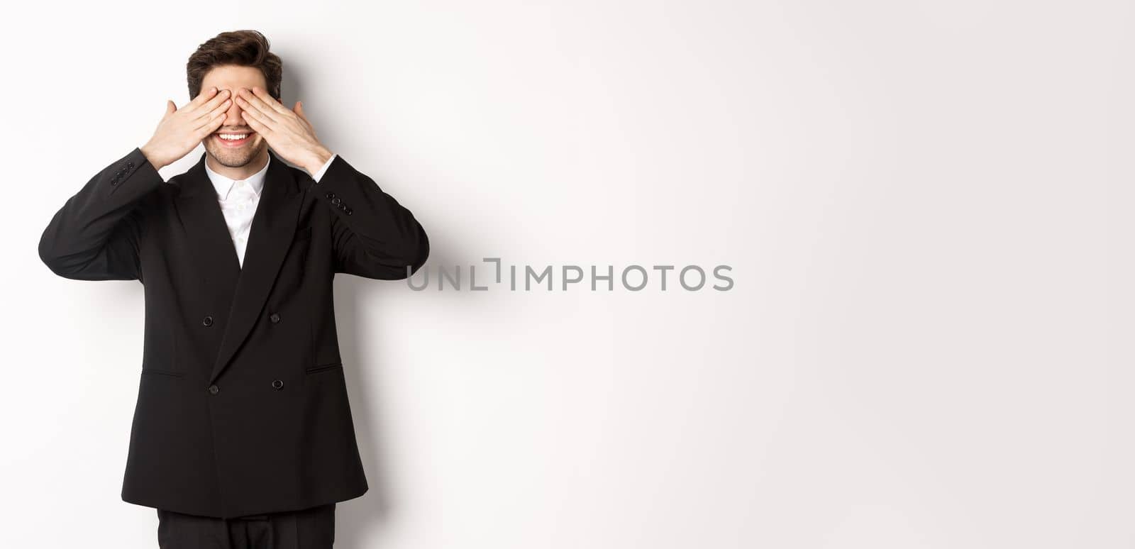 Image of handsome stylish man in black suit, waiting for christmas surprise, covering eyes with hands and smiling, anticipating presents, standing over white background.