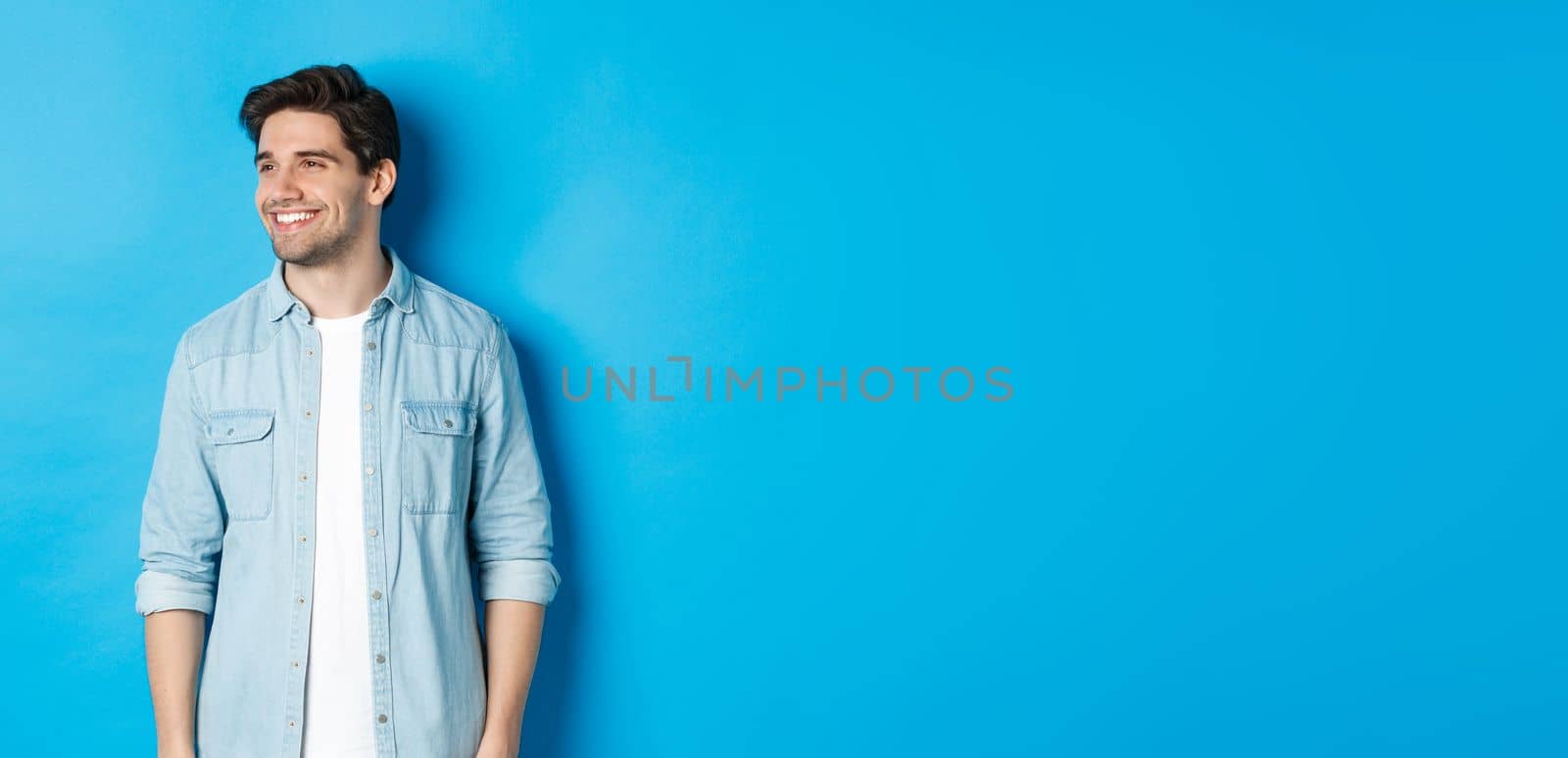 Handsome smiling adult man in casual outfit, smiling and looking left at promo offer, standing against blue background.