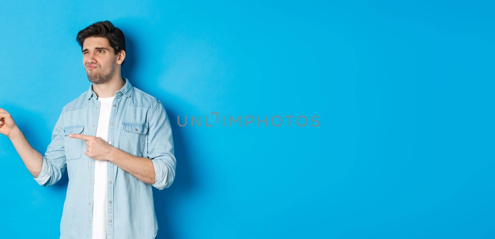 Doubtful adult man pointing fingers left at promotion and looking unsure, grimacing disappointed, standing against blue background.