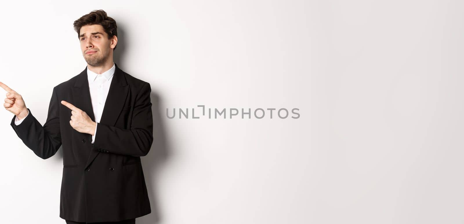Image of upset and disappointed handsome guy in formal suit, pointing and looking left with sad face, standing over white background by Benzoix