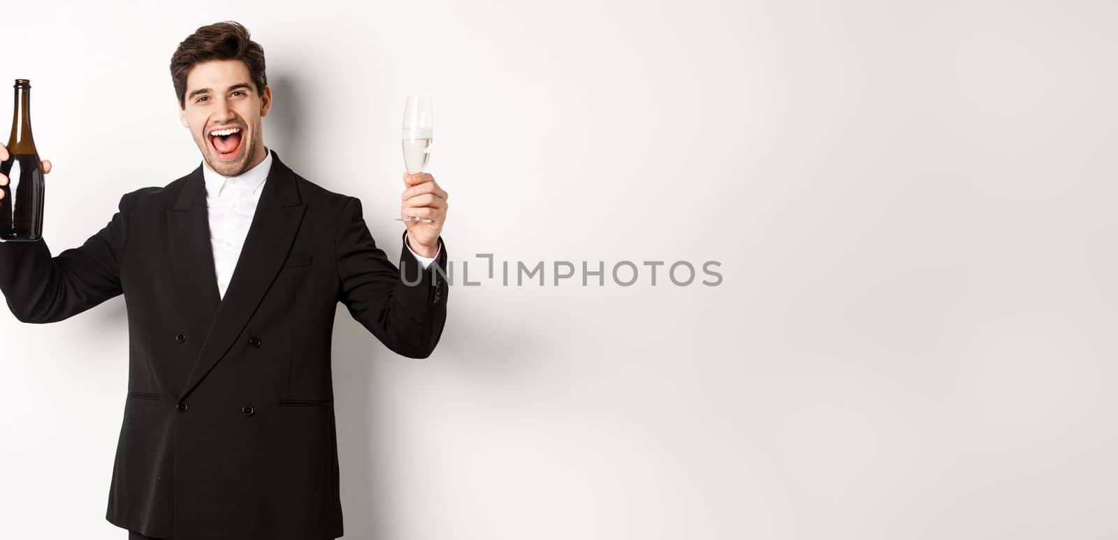 Concept of holidays, party and celebration. Handsome man in trendy suit having fun, holding bottle and glass of champagne, standing over white background by Benzoix