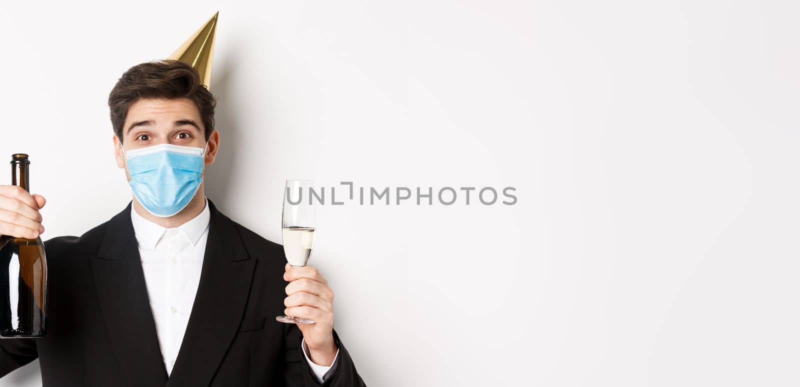 Concept of party during covid-19. Close-up of handsome man in suit, funny hat and medical mask, holding bottle of champagne, celebrating new year during coronavirus.