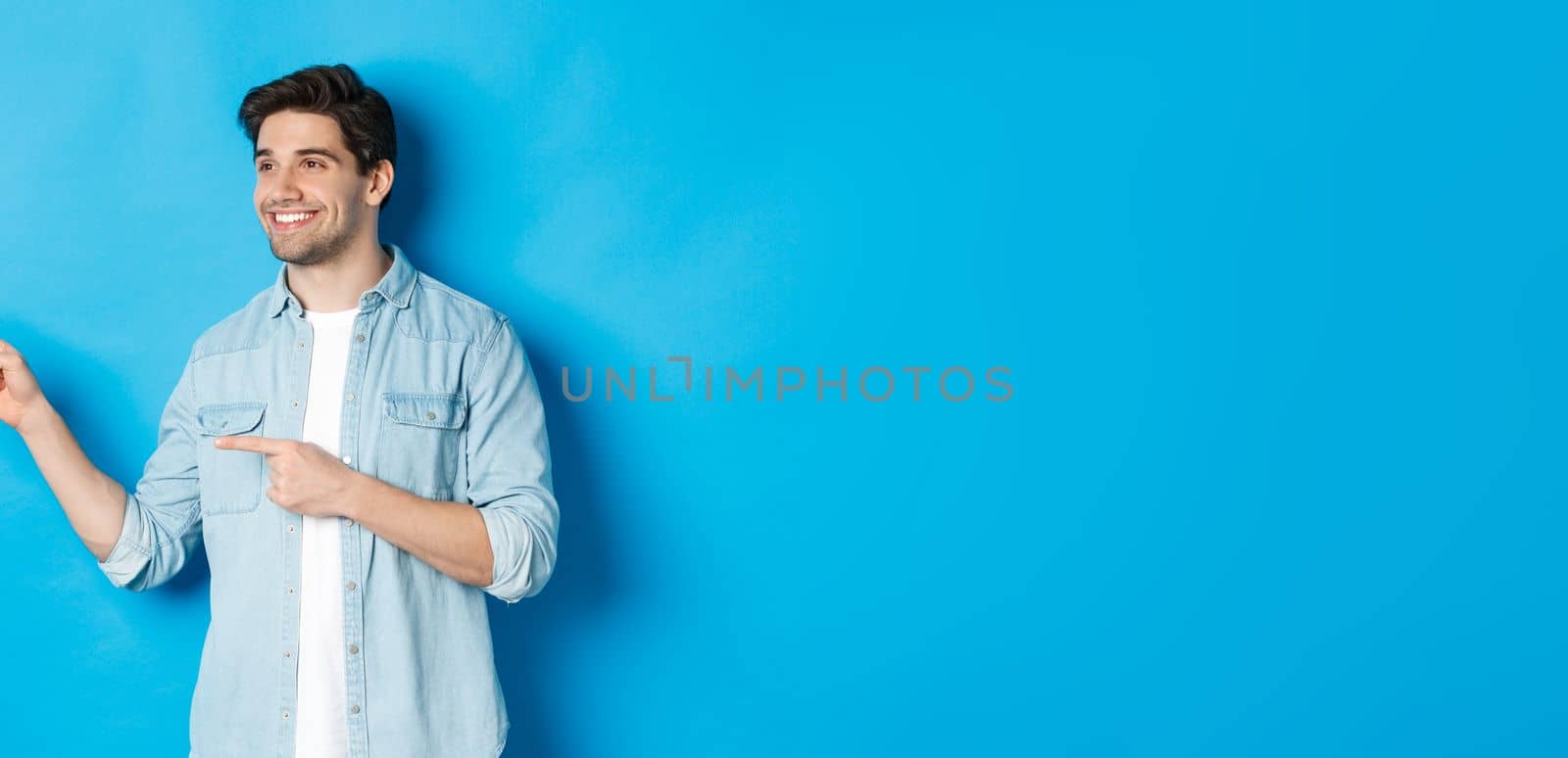 Handsome adult man introduce product, looking and pointing fingers left, promoting something against blue background by Benzoix