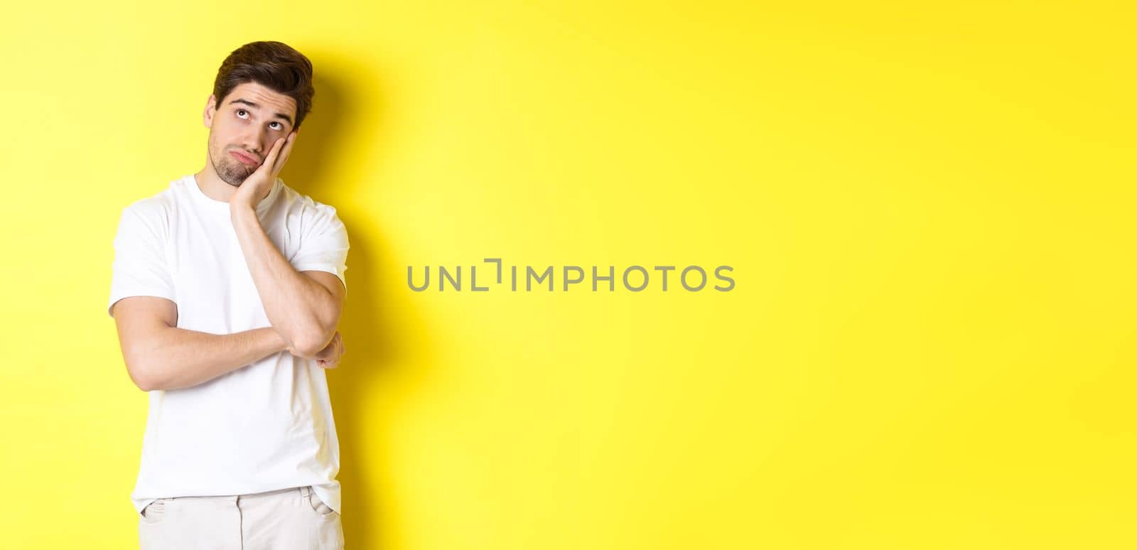 Bored and gloomy guy looking up, imaging things, standing over yellow background.