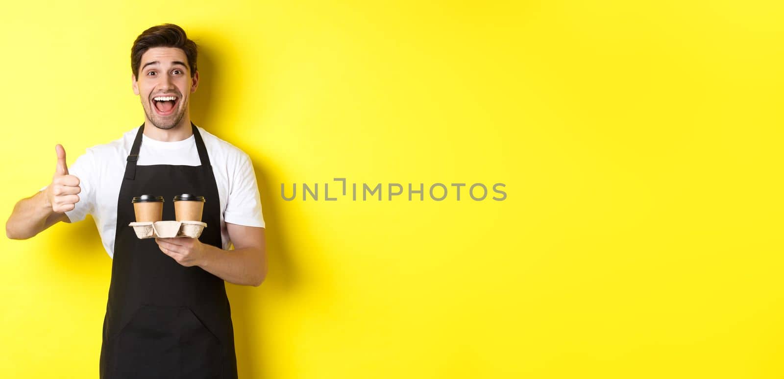 Cheerful barista recommend his cafe, holding takeaway coffee cups and showing thumbs up, standing against yellow background.