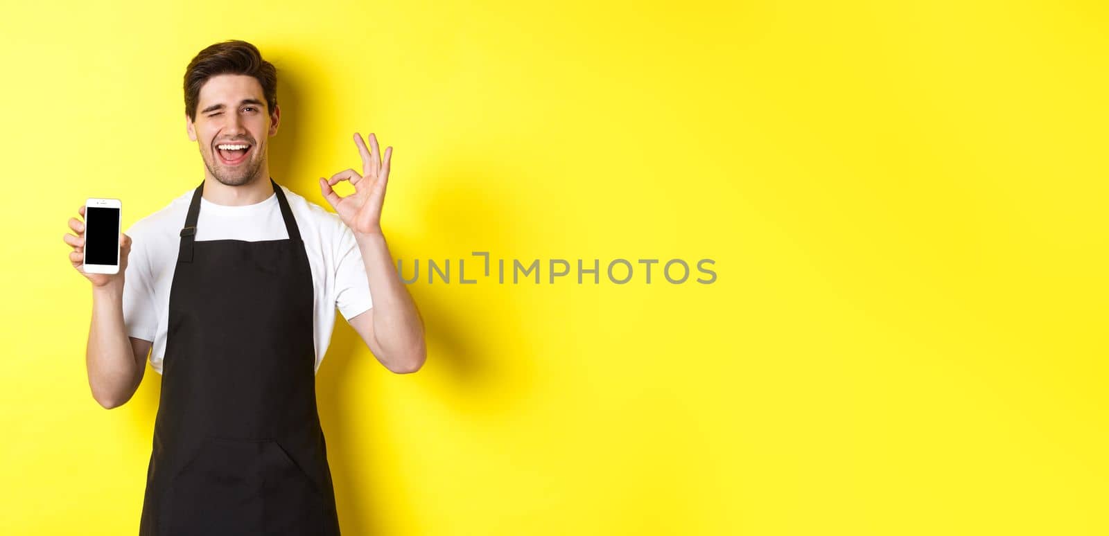 Handsome coffee shop worker showing ok sign and smartphone screen, recommending application, standing over yellow background.