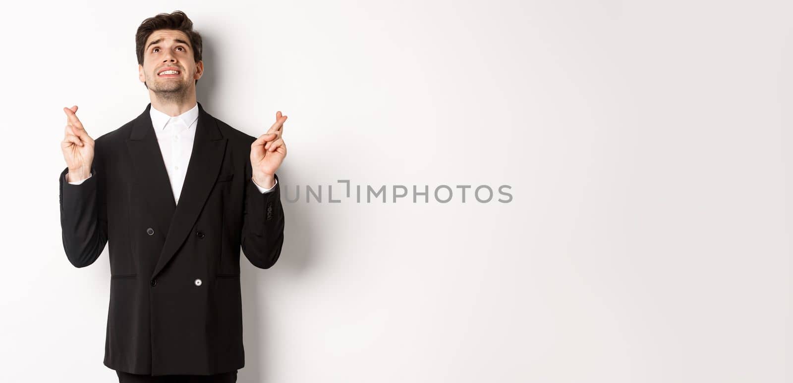 Portrait of tensed and worried handsome businessman, crossing fingers and looking up, begging god, making a wish, standing against white background in black suit.