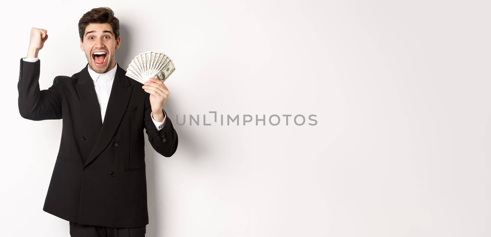 Portrait of handsome businessman in black suit, winning money and rejoicing, raising hand up with excitement, standing against white background.