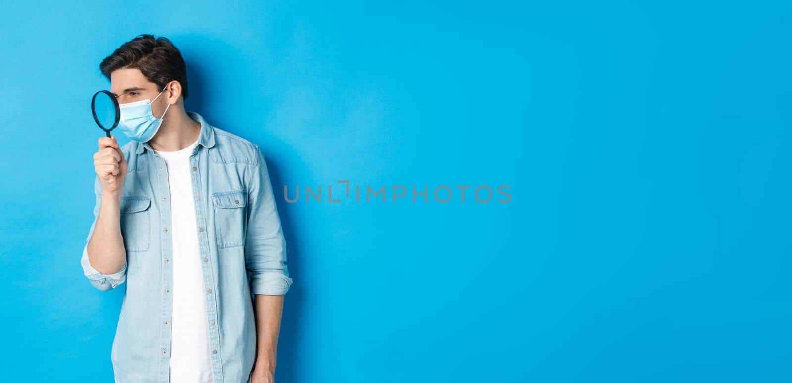 Concept of coronavirus, social distancing and pandemic. Man in medical mask searching for something, looking left through magnifying glass, studying copy space, blue background.