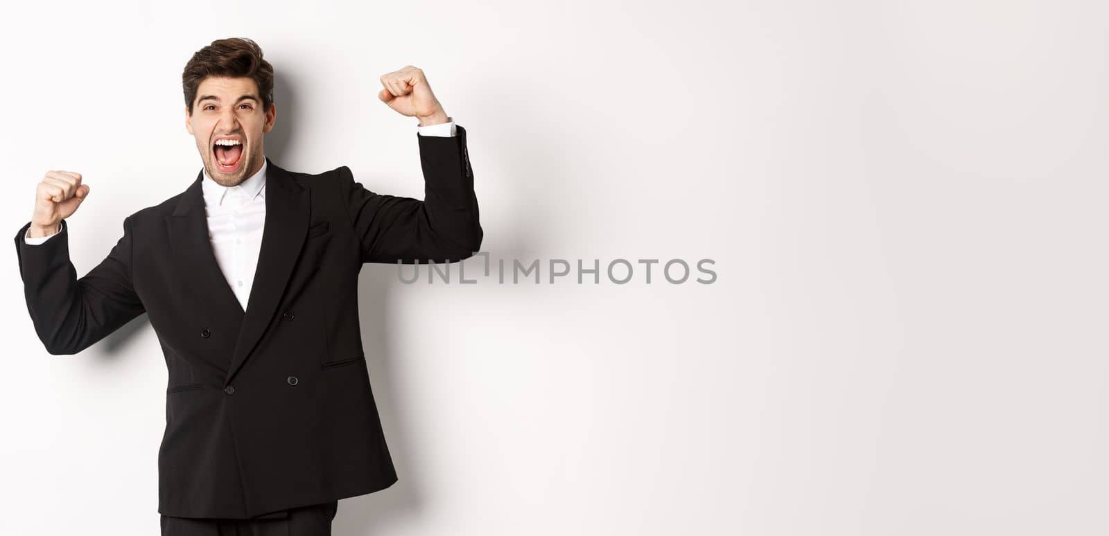 Portrait of successful businessman in black suit, become champion, raising hands up and shouting yes, triumphing and celebrating victory, standing against white background.